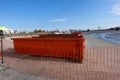 Two orange dumpsters at a construction site on a wet sandy beach near an orange mesh fence