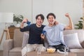 Two overjoyed young Asian male friends raised their hands, held game controllers, looked at the TV screen, and Royalty Free Stock Photo