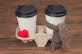 Two paper cups of coffee with knit heart and scarf on wooden background.