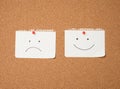 Two paper leaves with a smile and a sad emotion attached with a button on a brown board Royalty Free Stock Photo
