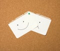 Two paper leaves with a smile and a sad emotion attached with a button on a brown board Royalty Free Stock Photo