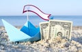 Two paper one dollars bills half buried in sand, blue paper boat Royalty Free Stock Photo