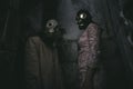 two people in gas masks stand against a wall in a small dark room