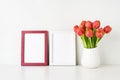 Two picture frame mockups and red tulips in a white vase on a white table by the wall. Copy space Royalty Free Stock Photo