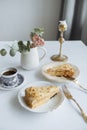 Two pieces of cake on white plates golden forks a vase with a flower and coffee and a golden candlestick on a white table