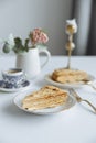 Two pieces of cake on white plates golden forks a vase with a flower and coffee and a golden candlestick on a white table