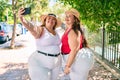 Two plus size overweight sisters twins women smiling taking a selfie picture with the phone outdoors on a sunny day