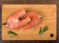 Two portioned salmon steaks and two sprigs of rosemary on a cutting board over a wooden background.