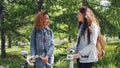 Two pretty female bicyclers with backpacks are talking standing in the park holding bikes. Pan shot of beautiful city