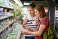 Two pretty female sisters go shopping together, stands in grocer`s shop, select fresh milk in paper container, read label, carry r Royalty Free Stock Photo