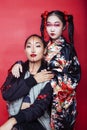 Two pretty geisha girls friends: modern asian woman and traditional wearing kimono posing cheerful on red background Royalty Free Stock Photo