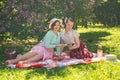 Two pretty girls friends sitting on the red blanket on the green grass and have summer picnic. happy woman having rest and fun on