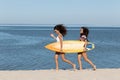 Two pretty girls are walking along the sandy beach near the sea and holding together a yellow surfboard in their hands Royalty Free Stock Photo