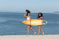 Two pretty girls are walking along the sandy beach near the sea and holding together a yellow surfboard in their hands Royalty Free Stock Photo