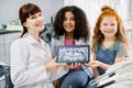 Two pretty smiling multiethnic teen girls, sitting on chair and looking at camera, while holding x-ray scan image of