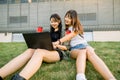 Two pretty young asian girls wearing trendy casual clothes, sitting on the green grass with laptop computer, drinking Royalty Free Stock Photo