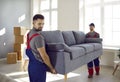 Two professional relocation service workers in overalls move sofa in customer& x27;s apartment.