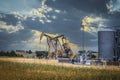 Two pump jacks and tanks and oil field equipment in fenced area in pasture under dramatic skies with houses on hill on horizon - Royalty Free Stock Photo