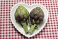 TWO PURPLE FRESH ARTICHOKE ON A HEART SHAPE WHITE PLATE ON RED