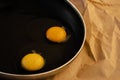 Two raw eggs with bright yellow yolks and transparent whites with a clots in a black frying pan on paper. The concept of cooking