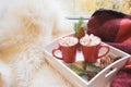 Two red cup of hot chocolate with marshmallow on white windowsill with furskin for rest. Holiday. Christmas morning.