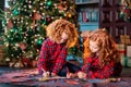 Two red-haired curly-haired girls write letter to Santa against the backdrop of Christmas tree and a decorated room