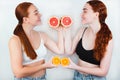 Two redheaded women holding two halves of grapefruit and two halves of orange in hands standing in front of each other on 
