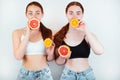 Two redheaded young women holding two halves of grapefruit and two halves of orange in hands standing on isolated white background