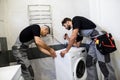 Two repairmen, workers in uniform working, examining washing mashine using flashlight in the bathroom. Repair service