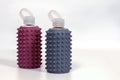 Two reusable glass bottles for water in dusty pink and gray rubber cases, with many spikes and plastic tops, comfortable for finge