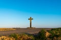 Two rural roads intersect at a big old Christian stone cross in front of a clear blue sky at the beginning sunset