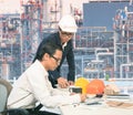 Two of same engineer working on table against exterior of oil refinery p
