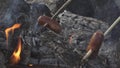 Two sausages attached to sticks are baking on flame of campfire