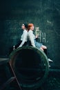 Two schoolgirls sitting side by side on a concrete ring near an old concrete wall. Industrialization, street culture, and urban