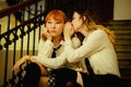 Two schoolgirls in uniform are sitting on the school stairs. The cute blonde is whispering her secret with her friend, a ginger Royalty Free Stock Photo