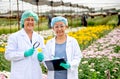 Two scientist women with lab gown and hair cover stand in front of multi-color flowers. They are also smiling with the concept of