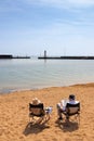 Two senior adults sat in portable chairs on a sandy beach reading books