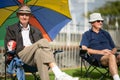 Two senior men , one under colourful sun umbrella sitting on grass during 60th National Jazz Festival in city