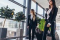 Two serious young female colleagues walking through airport passageway carrying suitcases Royalty Free Stock Photo