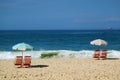 Two set of red beach chairs and a blue beach parasol on the sandy beach facing the crashing waves and vivid blue ocean
