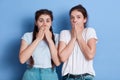 Two shocked women dress whit casual t shirts posing against blue wall covering their mouths, standing with shocked facial Royalty Free Stock Photo