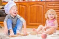 Two siblings - boy and girl - in chef`s hats sitting on the kitchen floor soiled with flour, playing with food, making mess and ha