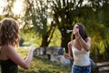 Two sisters, blowing soap bubbles at each other in park, laughing, smiling, Young pretty girls, wearing jeans shorts, green beige Royalty Free Stock Photo