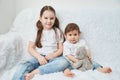 Two children, sisters sit on a white sofa in white t-shirts and blue jeans. Soft plush bear