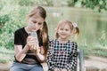 Two sisters sitting on bench in spring summer park near the pond and watching cartoons on smartphone. Wireless technologies Royalty Free Stock Photo