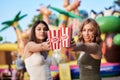 Two sisters, standing is front of attraction in theme park, holding popcorn in paper box, smiling, having fun. Pretty girls.