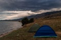Two small blue and green tents stand on grassy shore of bay of Lake Baikal, seas with waves, among trees and mountains