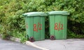 Two small green plastic mobile dumpsters are parked near the bushes