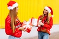 Two smiling beautiful young women in red sweaters and santa claus hats give each other christmas gifts while standing against Royalty Free Stock Photo
