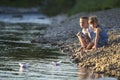 Two smiling blond children, boy and girl playing with white paper boats on river bank on bright summer blurred blue background. J Royalty Free Stock Photo
